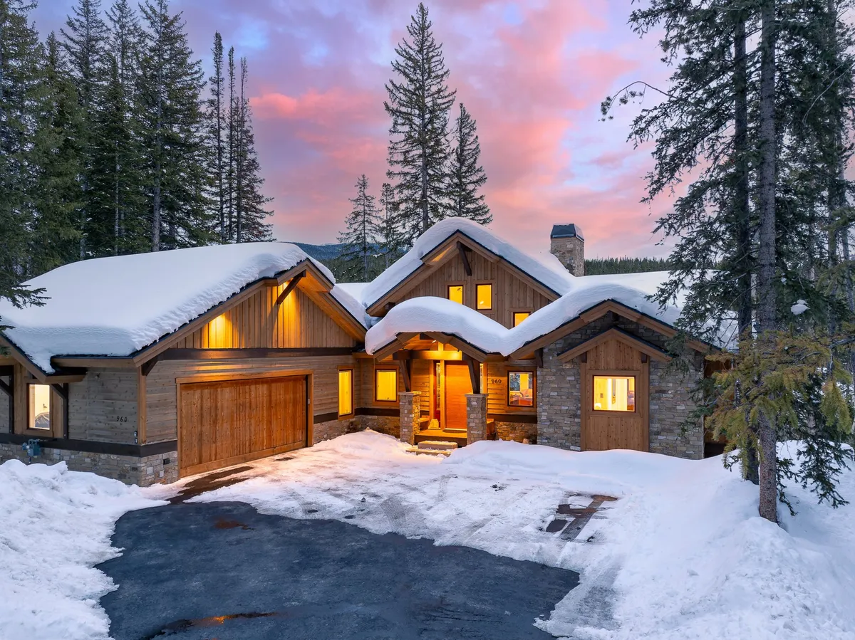 This image captures the exterior of the home, showcasing its warm glow from the windows, beautiful stone and wood architecture, and a snow-covered landscape surrounding the property. The inviting ambiance hints at a welcoming retreat ideal for gathering with family and friends.