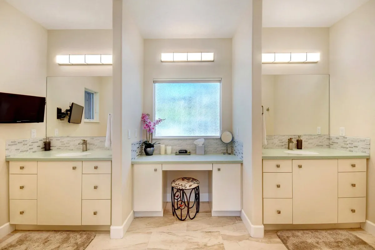 A modern double vanity bathroom bathed in natural light with elegant mirrors, a decorative stool, and clean lines, evoking a sense of refreshment and relaxation. A TV is visible, adding a contemporary touch.
