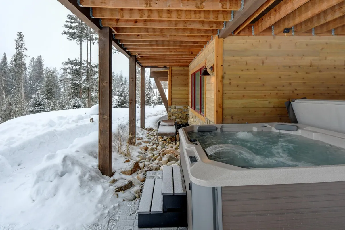 This image captures a steaming outdoor hot tub nestled among snow-covered ground and surrounded by trees, with rustic wooden beams accenting the setting. The tranquil winter scene invites relaxation and showcases the kind of private, resort-style amenity described in the section.