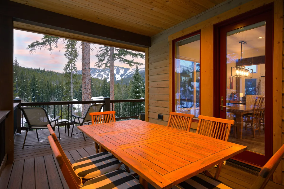 A wooden outdoor dining table is set on a spacious deck or patio, framed by mountain views and shaded by trees. The setup is perfect for alfresco dining, family gatherings, or simply enjoying nature with loved ones.