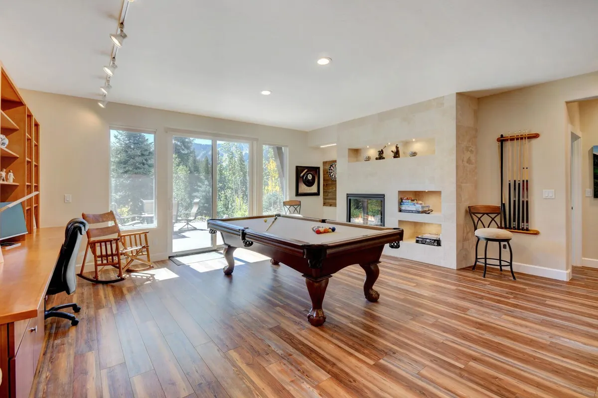 This cozy interior scene highlights a pool table adjacent to a fireplace and large windows, with a visible bookshelf in the background. The room invites both social game nights and quiet reading, blending recreation with relaxation.