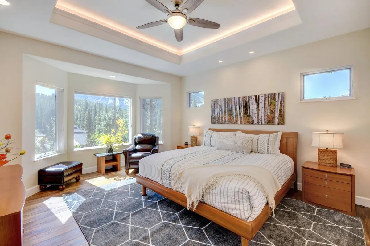 A king-sized bed sits under large windows with natural light streaming in, complemented by warm wooden furniture and tasteful artwork on the wall. The setting is serene and spacious, ideal for restful nights and private moments.