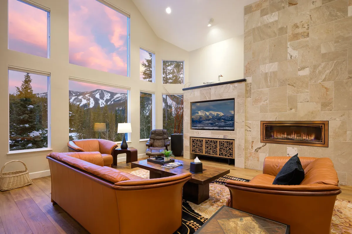A warm and welcoming living room featuring expansive floor-to-ceiling windows that frame breathtaking Colorado mountain views. The space is anchored by a gas fireplace, with plush leather furnishings arranged for relaxation and socializing, and a 65-inch Smart TV positioned for movie or game night enjoyment. The room glows with natural light, emphasizing the inviting atmosphere ideal for gatherings.
