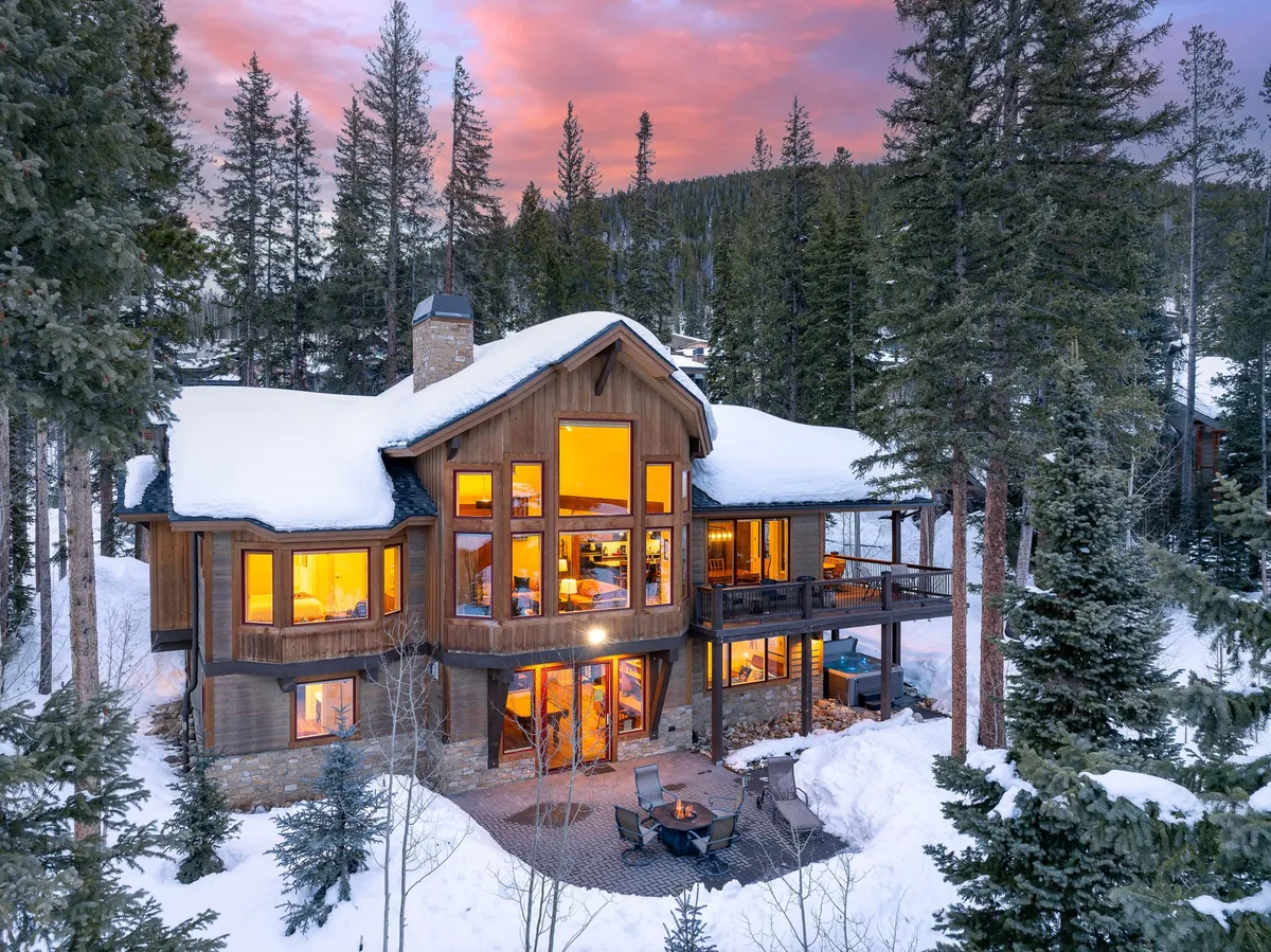 The image shows a luxurious mountain home warmly lit from within, surrounded by snow-dusted pines. The spacious deck features a cozy fire pit with inviting outdoor seating, perfect for gathering and soaking in panoramic winter views.