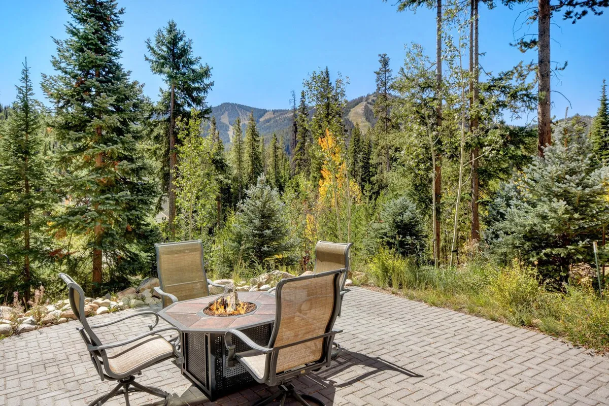 An inviting outdoor fire pit takes center stage, surrounded by comfortable seating and tall trees, with a majestic mountain backdrop. The scene is set during sunset, with glowing firelight reflecting off the surroundings, evoking warmth and togetherness.