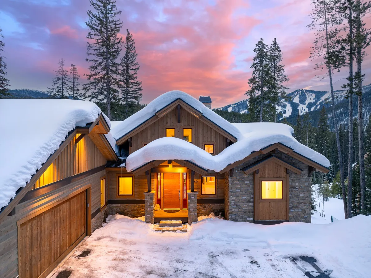 A welcoming exterior view of the home, featuring a snow-covered roof, warmly lit windows, and a dramatic mountain backdrop. People can imagine gathering at the entrance or heading out for winter adventures, while the inviting atmosphere hints at cozy evenings inside.