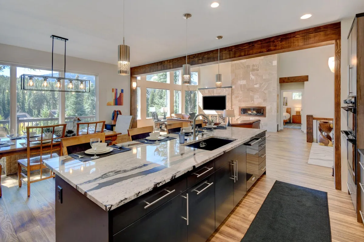 A spacious, modern kitchen and dining area seamlessly connected, with a prominent kitchen island, large windows streaming in light, a nearby fireplace, and a glimpse of the living space. The setup invites communal cooking, dining, and entertaining, all while enjoying scenic views of the mountains.